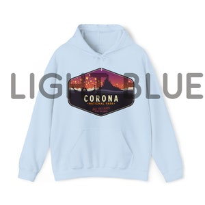 Puede incluir: Sudadera con capucha azul claro con un gr&aacute;fico frontal que presenta una escena nocturna con un castillo, globos aerost&aacute;ticos y el texto "CORONA NATIONAL PARK". La sudadera tiene un bolsillo delantero y una capucha con cord&oacute;n.