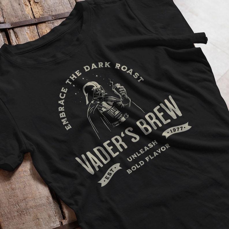 May include: Black t-shirt with a graphic of Darth Vader holding a cup of coffee. The text reads "Embrace the Dark Roast Vader's Brew Est. 1977 Unleash Bold Flavor".
