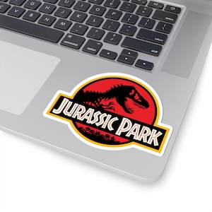 May include: A sticker featuring the "Jurassic Park" logo. The design includes a black dinosaur silhouette against a red circle, with the words "JURASSIC PARK" in a bold, yellow-outlined font. The sticker is placed on a laptop.
