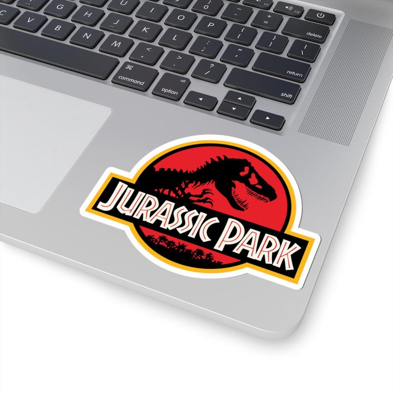 May include: A sticker featuring the "Jurassic Park" logo. The design includes a black dinosaur silhouette against a red circle, with the words "JURASSIC PARK" in a bold, yellow-outlined font. The sticker is placed on a laptop.