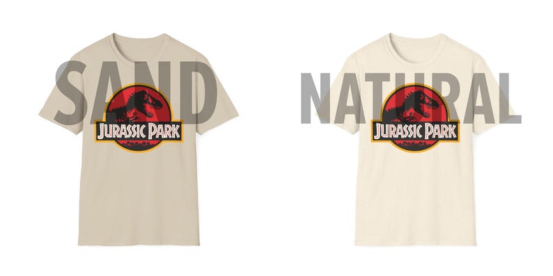 May include: Two t-shirts, one sand-colored and one natural-colored, each featuring the Jurassic Park logo. The logo includes a red circle with a dinosaur silhouette, the words "JURASSIC PARK" in white, and a yellow border.