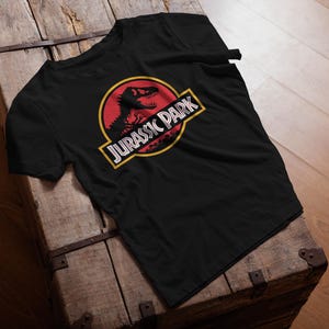Black unisex T-shirt with the classic Jurassic Park red and yellow dinosaur logo, retro 90s nostalgia graphic tee for men and women.