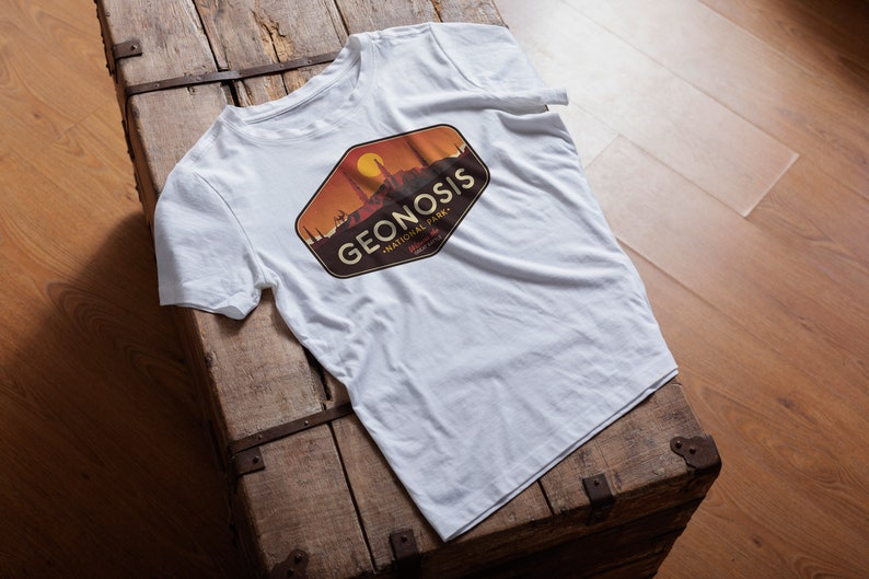 May include: White t-shirt featuring a brown, orange and yellow graphic design of a cityscape with the text "GEONOSIS - NATIONAL PARK" in a vintage style.