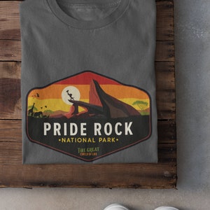 May include: A dark gray t-shirt with a vintage-style graphic of Pride Rock from The Lion King. The graphic features a sunset, a lion, and the text "Pride Rock National Park. The Great Circle of Life."