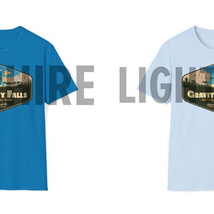 K&ouml;nnte beinhalten: Zwei T-Shirts mit einem grafischen Design eines Retro-Nationalpark-Logos. Das Logo zeigt eine Bergkette, einen See und den Text "Gravity Falls National Park". Das T-Shirt links ist saphiblau, das T-Shirt rechts ist hellblau.