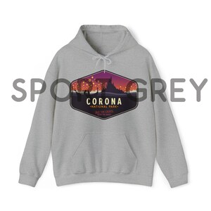 Puede incluir: Una sudadera con capucha gris claro con un bolsillo delantero y una capucha con cord&oacute;n. La sudadera presenta un gr&aacute;fico de un castillo y linternas flotantes con el texto "CORONA NATIONAL PARK" y "SEE THE LIGHTS, ENJOY THE SHOW".