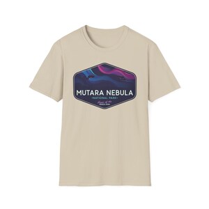 May include: A beige t-shirt featuring a graphic of the Mutara Nebula National Park. The graphic is a dark blue, purple, and pink design inside a dark blue hexagon. The text "MUTARA NEBULA" and "NATIONAL PARK" are also included.