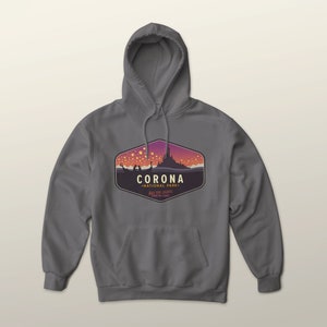 Puede incluir: Una sudadera con capucha gris con un bolsillo delantero y un cord&oacute;n. La parte delantera presenta un gr&aacute;fico de un castillo y linternas flotantes, con el texto "CORONA NATIONAL PARK" y "SEE THE LIGHTS".