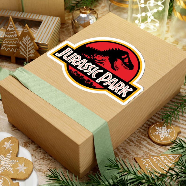May include: A sticker featuring the "Jurassic Park" logo, with a red circle and black dinosaur silhouette. The sticker is on a light brown gift box with a green ribbon. Christmas decorations are in the background.