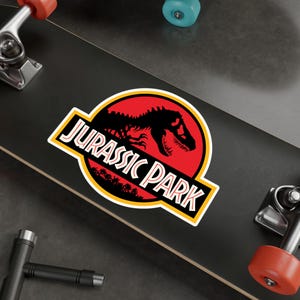 May include: A sticker featuring the Jurassic Park logo. The logo includes a black dinosaur silhouette inside a red circle, with the words "JURASSIC PARK" in white and yellow lettering. The sticker is on a black surface.
