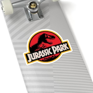 May include: A sticker featuring the "Jurassic Park" logo. The design includes a black dinosaur silhouette against a red background within a yellow-bordered circle. The words "JURASSIC PARK" are in a yellow-bordered black rectangle.