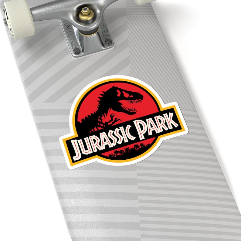 May include: A sticker featuring the "Jurassic Park" logo. The design includes a black dinosaur silhouette against a red background within a yellow-bordered circle. The words "JURASSIC PARK" are in a yellow-bordered black rectangle.