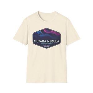 May include: Cream-colored t-shirt featuring a graphic of the Mutara Nebula National Park. The design includes a dark blue and purple nebula illustration within a dark blue hexagon shape. The text "MUTARA NEBULA" and "NATIONAL PARK" are also displayed.