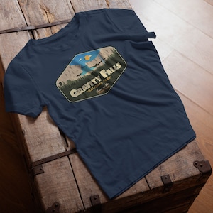K&ouml;nnte beinhalten: Ein marineblaues T-Shirt mit einer Grafik im Vintage-Stil eines Nationalpark-Logos. Das Logo zeigt eine Bergkette, einen See und eine fliegende Untertasse. Der Text "Gravity Falls National Park" ist in einem gelben Banner geschrieben.