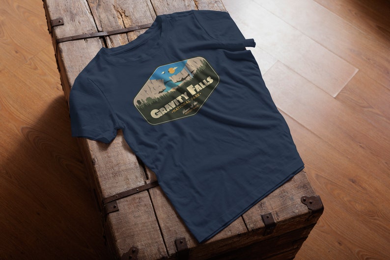 K&ouml;nnte beinhalten: Ein marineblaues T-Shirt mit einer Grafik im Vintage-Stil eines Nationalpark-Logos. Das Logo zeigt eine Bergkette, einen See und eine fliegende Untertasse. Der Text "Gravity Falls National Park" ist in einem gelben Banner geschrieben.