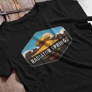 以下が含まれることがあります： 黒いTシャツ。砂漠の風景のグラフィックデザインで、「RADIATOR SPRINGS NATIONAL PARK」の文字が白で描かれています。デザインには太陽、青い空、岩の形が含まれています。Tシャツは柔らかい素材で作られています。