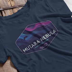 May include: Navy blue t-shirt featuring a graphic of the Mutara Nebula National Park. The design includes a stylized nebula with two spacecraft and the text "MUTARA NEBULA NATIONAL PARK Home of the Genesis Spark."