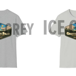 K&ouml;nnte beinhalten: Zwei T-Shirts in verschiedenen Graut&ouml;nen, beide mit einem Grafikdruck eines Retro-Nationalpark-Logos mit dem Text "Gravity Falls National Park" und einer Illustration einer Bergkette und eines Sees. Das Logo hat eine sechseckige Form mit gelbem Rand und braunem Hintergrund.