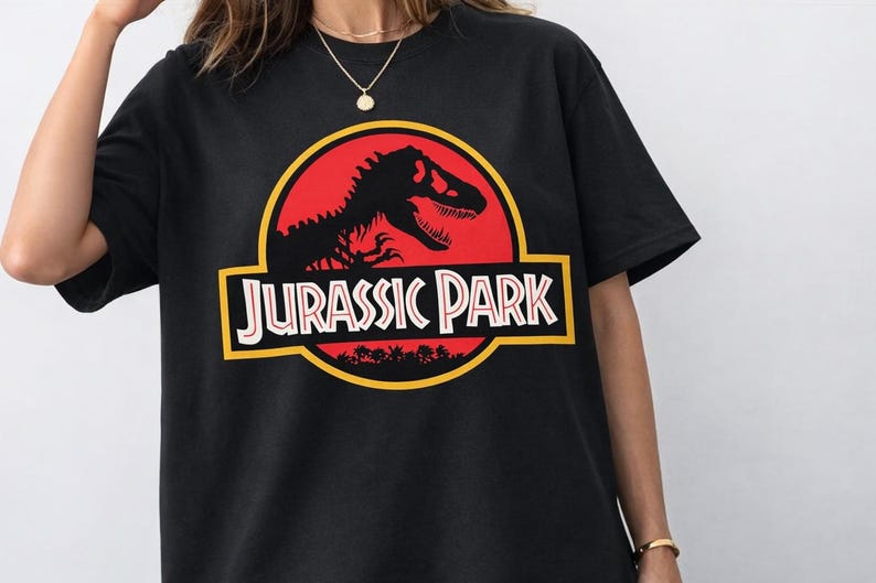 May include: Black t-shirt featuring the Jurassic Park logo. The logo includes a red circle with a dinosaur silhouette, the words "JURASSIC PARK" in white, and a yellow border. The shirt is short-sleeved.
