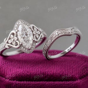 May include: A silver-toned engagement ring set featuring a marquise-cut center stone surrounded by a halo of small round stones. The band displays intricate floral engravings and heart-shaped accents, accompanied by a matching curved wedding band.