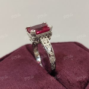 May include: A silver ring featuring a large, square-shaped red gemstone set in a filigree design. The ring is displayed on a red velvet background.