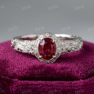 May include: A silver ring with an oval, deep red gemstone at the center, surrounded by smaller clear stones. The band features an intricate floral pattern. The ring is set on a dark purple velvet surface.