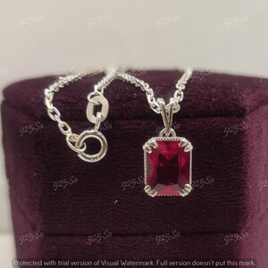 Puede incluir: Un collar de cadena de plata con un colgante que presenta una piedra preciosa roja en un engaste cuadrado. El collar se muestra en una caja de terciopelo burdeos.