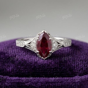 May include: A silver ring with a marquise-cut, deep red gemstone as the focal point. The ring features intricate detailing with smaller, clear gemstones and a decorative band. The ring is set on a purple velvet surface.