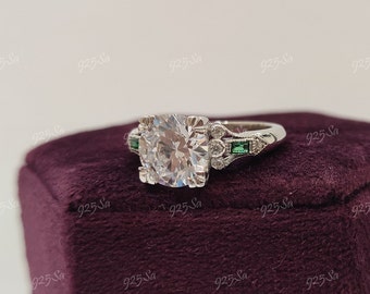 May include: A silver ring with a large, round, clear center stone and smaller, clear stones on the band. The band is decorated with green stones on either side of the center stone.