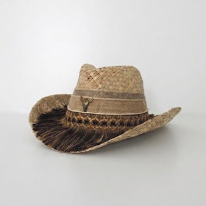 May include: A tan straw hat featuring a wide brim and a pinched crown. The hat is decorated with a textured beige band, a metallic bull skull emblem, and a dark brown fringe trim that encircles the base of the crown above the brim.