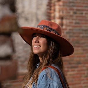 Può includere: Un cappello color ruggine a tesa larga con una fascia decorativa con un motivo blu e arancione. Il cappello ha una corona arrotondata e un'ampia tesa. Il cappello è indossato con una camicia di jeans e una borsa di cuoio marrone.