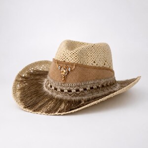 Op de afbeelding: Een strooien cowboyhoed met een beige jute band, versierd met een centraal goudkleurig stierenschedel-embleem ingelegd met heldere kristallen. De rand is afgewerkt met een brede, metallic bronzen franje en een geweven latwerkdetail.