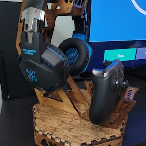 Pode incluir: Um suporte de madeira para fones de ouvido para jogos com um suporte de controlador correspondente. O suporte contém um fone de ouvido Speed Spider GS-G2000 preto e azul e um controlador preto. O suporte é feito de madeira marrom clara com um design geométrico.