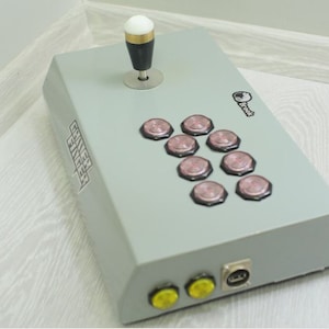 May include: A gray arcade game controller with a joystick and eight pink buttons. The controller has a key graphic and the text "Gamer Finger". It has two yellow buttons and a USB port.