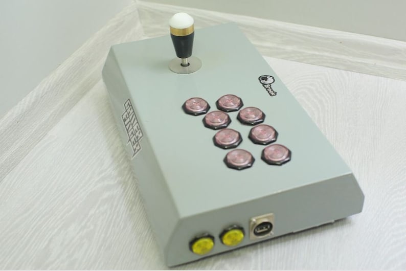 May include: A gray arcade game controller with a joystick and eight pink buttons. The controller has a key graphic and the text "Gamer Finger". It has two yellow buttons and a USB port.