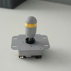 Può includere: Un joystick arcade grigio con un'impugnatura cilindrica caratterizzata da una fascia color oro al centro. L'unità include una piastra di montaggio quadrata in metallo con quattro viti visibili e terminali elettrici alla base.