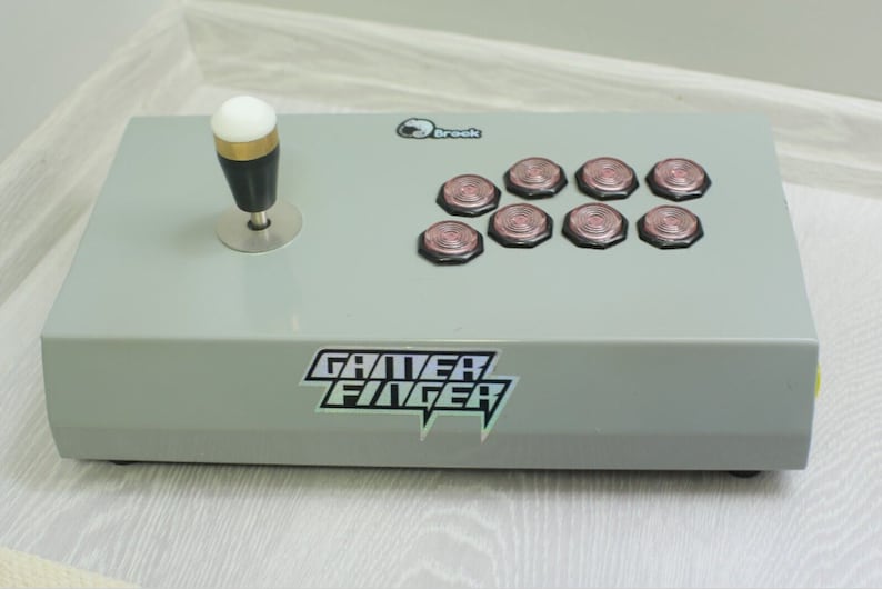 May include: A gray arcade controller with a joystick and eight buttons. The joystick has a white ball top, black shaft, and silver base. The buttons are pink and black. The text "GAMER FINGER" is visible on the front.