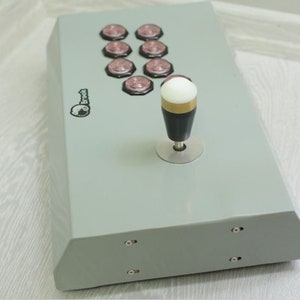 May include: A gray arcade game controller with eight pink buttons and a joystick. The joystick has a white ball top, a gold ring, and a black handle. The controller has the "Brook" logo.
