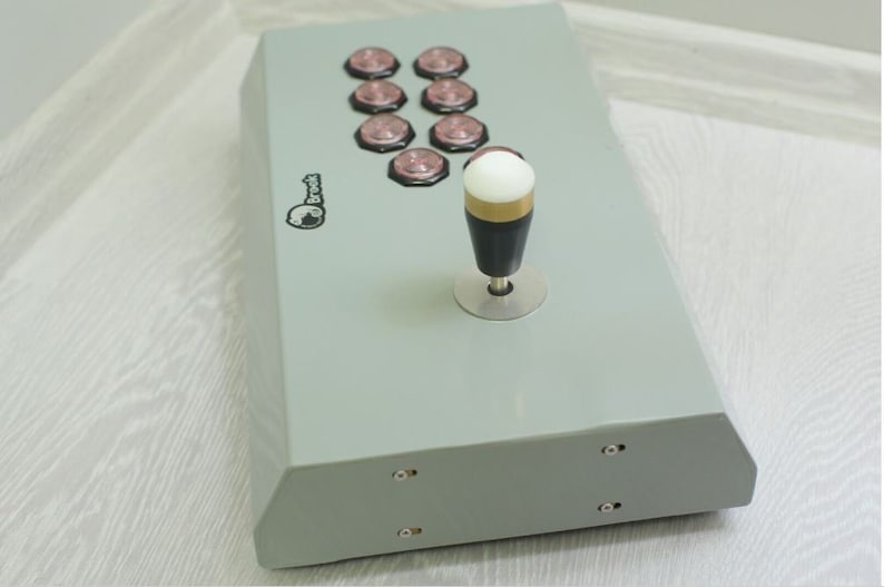 May include: A gray arcade game controller with eight pink buttons and a joystick. The joystick has a white ball top, a gold ring, and a black handle. The controller has the "Brook" logo.