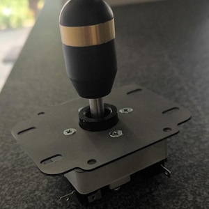 Può includere: Un controller joystick arcade dotato di un'impugnatura cilindrica nero opaco con una fascia orizzontale color oro. L'impugnatura è montata su una piastra di base rettangolare in metallo con quattro viti visibili e un alloggiamento del perno centrale nero.
