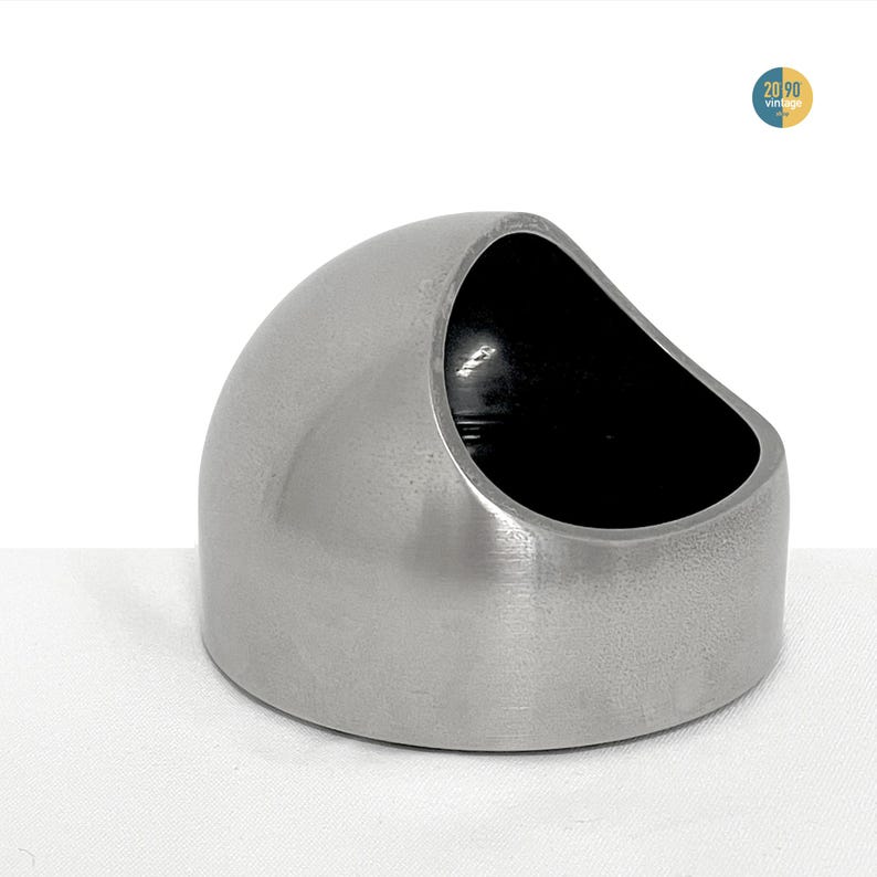 May include: A brushed stainless steel bowl with a dome-shaped exterior and a slanted, open top. The interior of the bowl is finished in a deep, solid black color. The object features a smooth, metallic surface with a circular base and a wide opening.