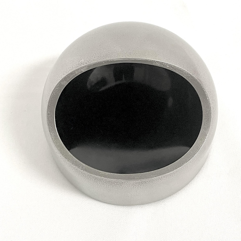 May include: A hemispherical object with a smooth, matte silver-toned exterior and a circular, glossy black inset on the flat face. The object features a clean, rounded dome shape that transitions into a flat base, presenting a high-contrast design.