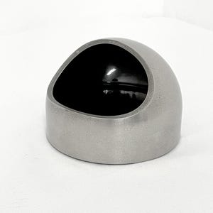 May include: A dome-shaped container with a matte silver exterior and a smooth, glossy black interior. The object features a wide, slanted circular opening that reveals the dark, reflective inner surface, creating a contrast between the metallic outer finish and the deep black hollow center.