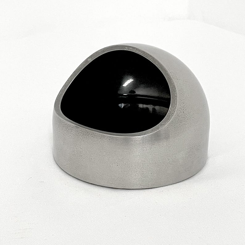 May include: A dome-shaped container with a matte silver exterior and a smooth, glossy black interior. The object features a wide, slanted circular opening that reveals the dark, reflective inner surface, creating a contrast between the metallic outer finish and the deep black hollow center.