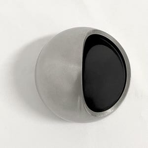May include: A spherical object with a smooth, matte silver exterior and a large, circular opening on one side. The interior of the opening is finished in a deep, solid black, creating a high-contrast visual effect against the metallic silver shell.