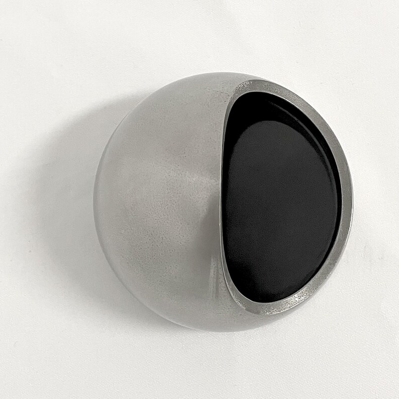 May include: A spherical object with a smooth, matte silver exterior and a large, circular opening on one side. The interior of the opening is finished in a deep, solid black, creating a high-contrast visual effect against the metallic silver shell.