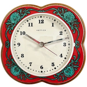 May include: A red and white clock with a floral design. The clock face has black numbers and hands. The clock is made by Hettich and says "Made in Germany" on the bottom.