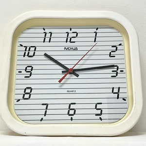 Könnte beinhalten: Eine weiße quadratische Wanduhr mit schwarzen Zahlen und Zeigern. Das Zifferblatt der Uhr hat einen weißen Hintergrund mit schwarzen Linien und das Wort "RADIUS" in schwarzen Buchstaben. Das Zifferblatt der Uhr hat auch das Wort "QUARTZ" in schwarzen Buchstaben. Die Uhr ist aus Kunststoff und hat einen weißen Rahmen.