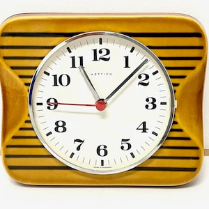 May include: A yellow ceramic clock with black stripes and a white face. The clock face has black numbers and a red second hand. The clock says "hettich" in the center.