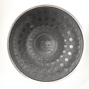 May include: A circular black rubber base with a metallic silver rim. The center features embossed text reading CINI&NILS, design OPI milano, modello depositato, and made in italy. The surface has a textured pattern of raised black circles.